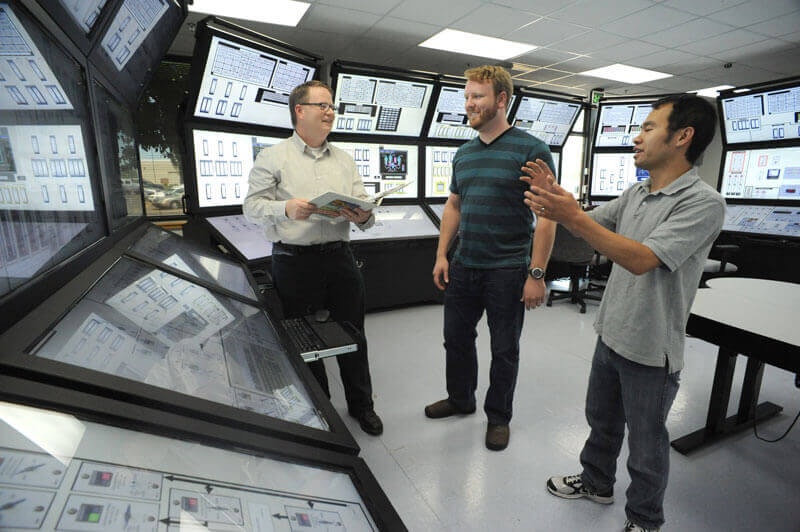 three men talking in a room filled with computer monitors and control screens